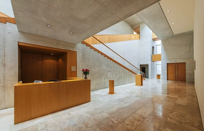 Offenes Foyer mit Empfangsbereich aus hellem Holz im Kontrast zu hellgrauem Betonboden und-Wänden