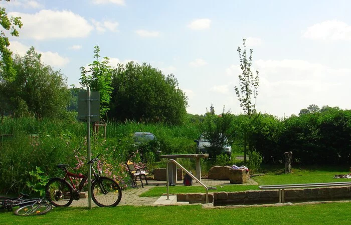 Wiese mitten im Grünen mit Tisch und Bänken und zwei Fahrräder. In der Mitte ein Kneipp Becken