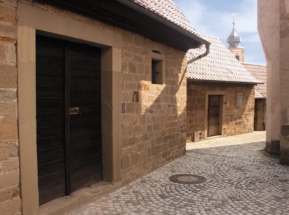 Eng bebaute Gasse mit kleinen nüchternen Steinhäusern, im Hintergrund Kirchturmspitze