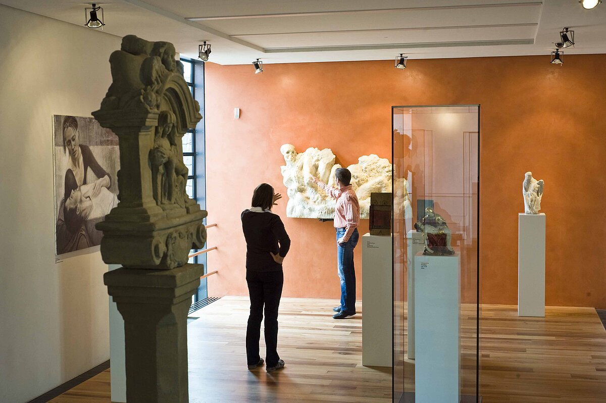 Raum mit Steinskulptur links, Glasvitrinen mit Ausstellungsstücken in der Mitte, an der hinteren orangen Wand hängt eine große helle Figur, Besucher schauen sich um