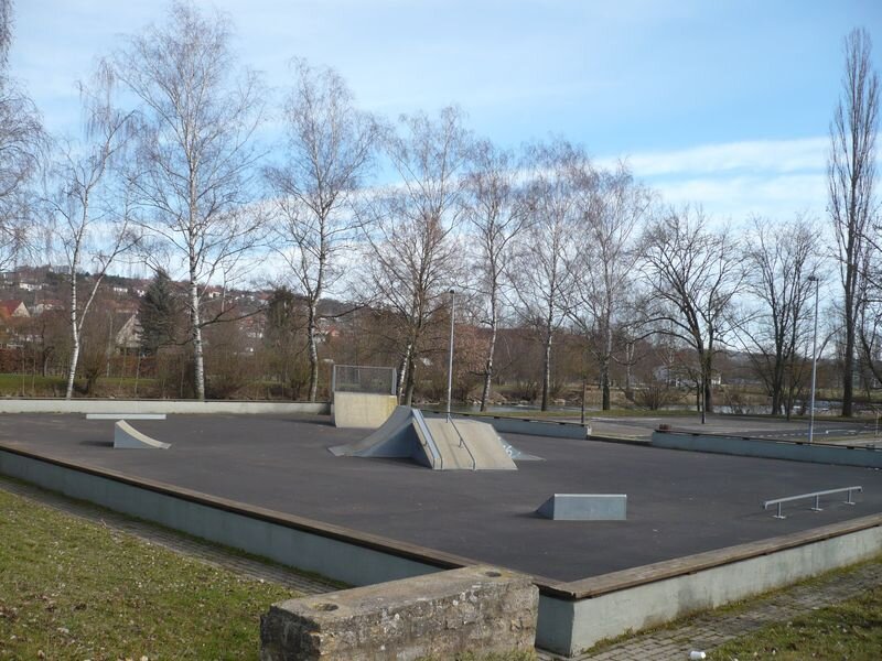 Menschenleerer eingefasster dunkler Platz  mit Hindernissen für Skater, eine Birkenreihe steht im Hintergrund