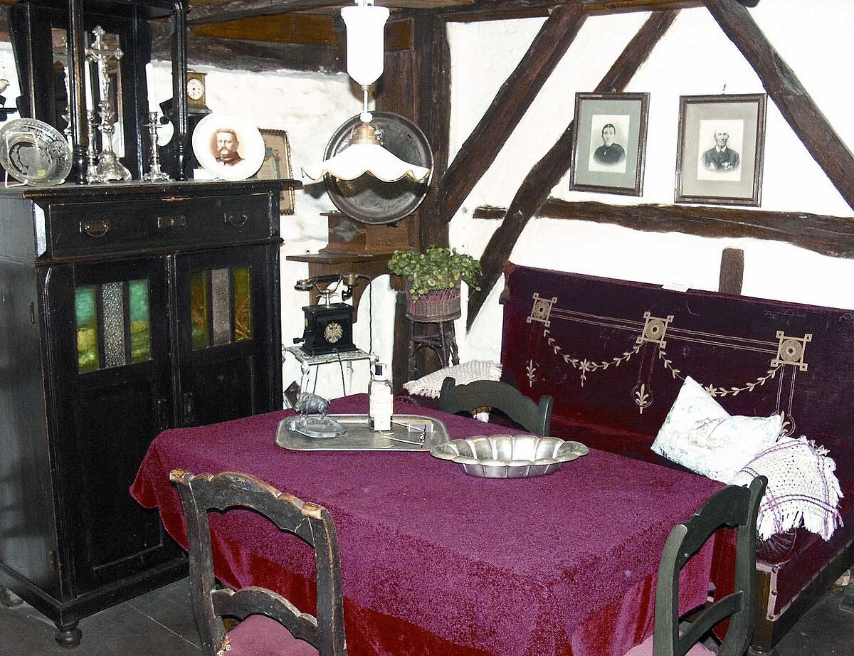 Museum in Gochsheim Innenraum, lilagedeckter Tisch mit 2 Stühlen, links dunkle Holzkommode, rechts an der Wand zwei Bilder und dunkle Holzleisten