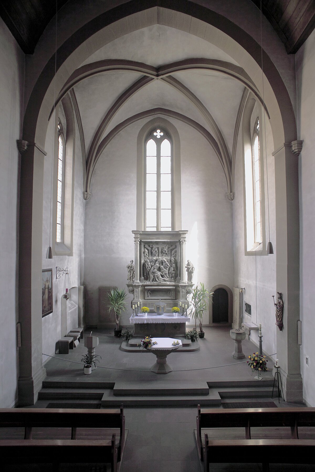 Blick über Mittelschiff in Altarraum einer Kirche, neogotische Fenster, vor mittlerem Fenster steht ein historischer Steinaltar