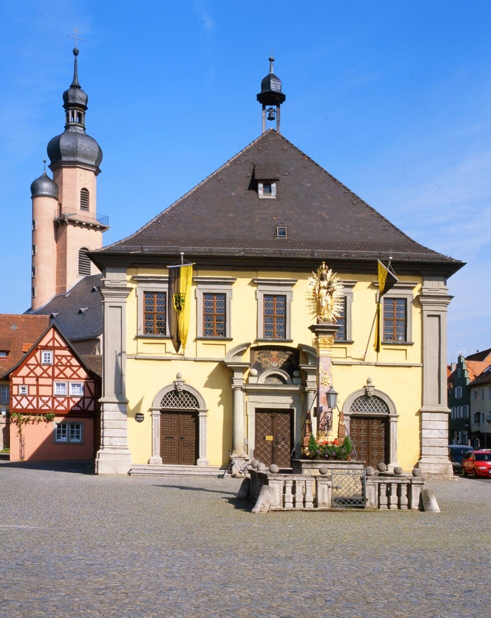 Gelbes barockes Haus mit schwarzem Dach, links dahinter zwei Kirchtürme aneinandergebaut in rosa