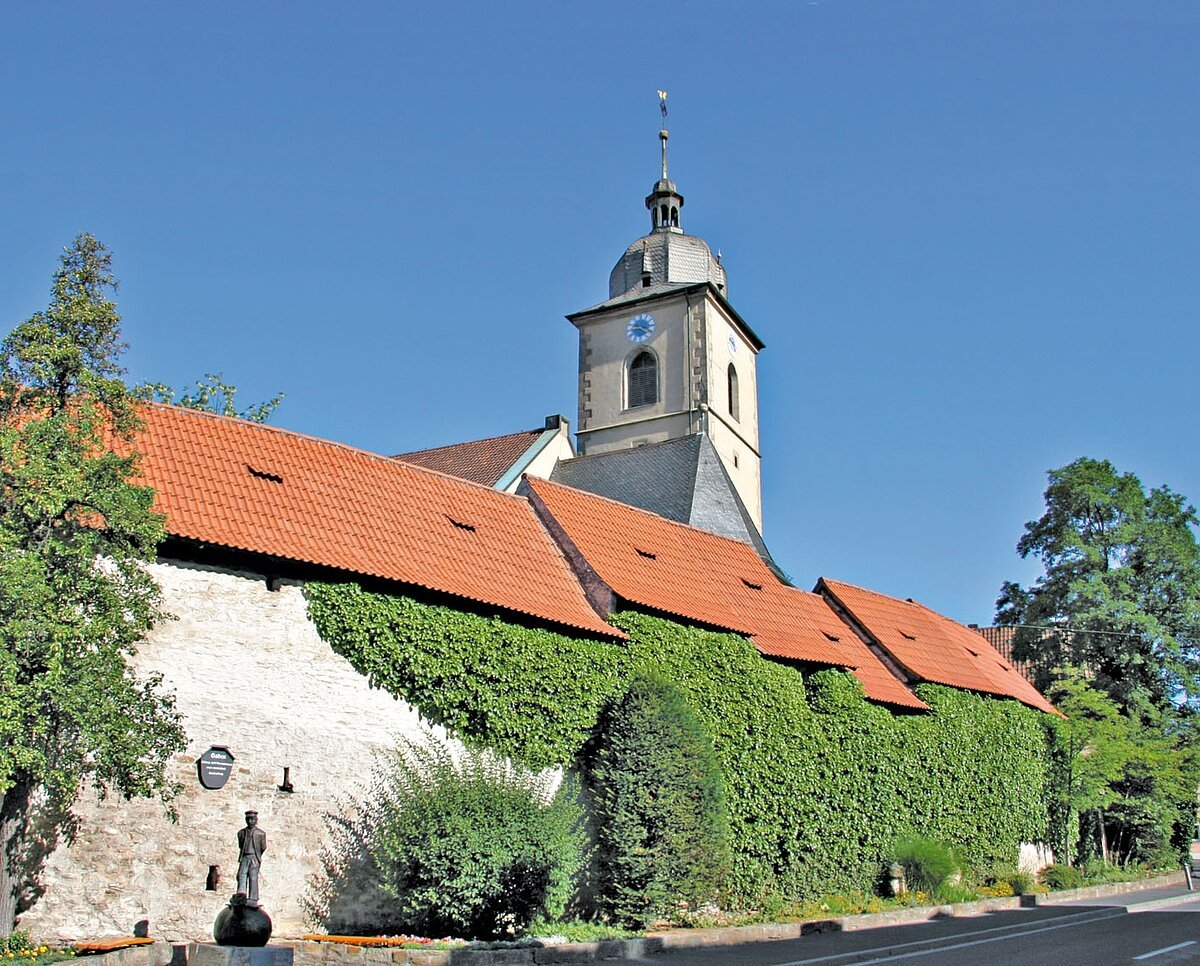 Kirchturm mit langgezogener Häuserreihe ohne Fenster mit grünbewachsenem Mauerwerk