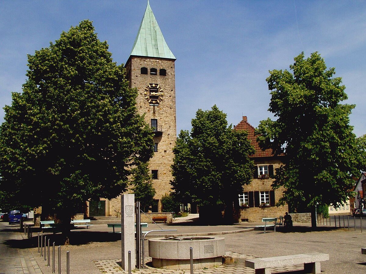 Moderner Kirchturm mit Uhr, sie steht an einem Platz Haus und Bäumen