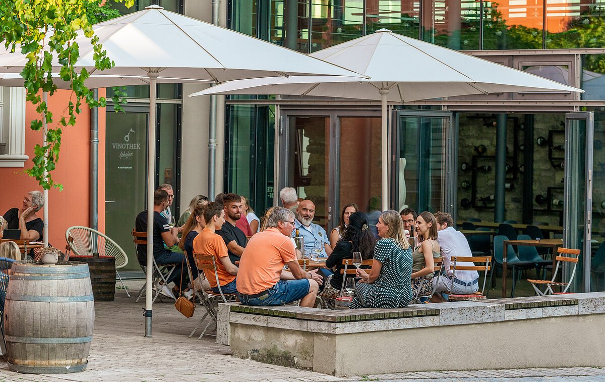 Menschen sitzen unter Sonnenschirmen vor der Vinothek Iphofen und genießen Wein und eine Brotzeit