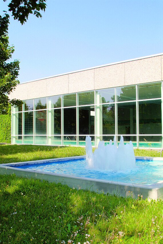Außenaufnahme: Gebäude mit Glasfront, grünem Rasen davor und kleinem Becken mit Springbrunnen in der Mitte