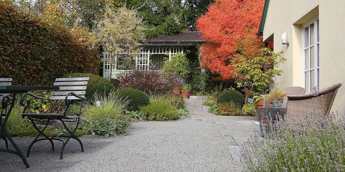 Gartenanlage mit Eingang und Weg , Sitzgelegenheit links und strahlendem orangenen Strauch hinter dem Haus
