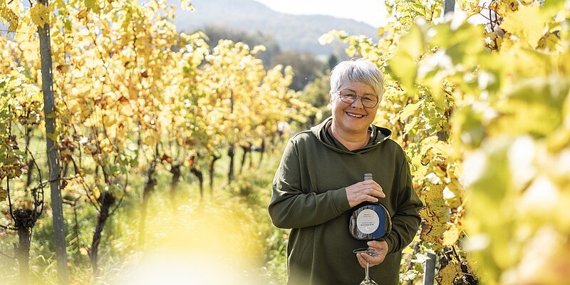 Winzerin Barbara Baumann Winzerin Barbara Baumann vom Weingut Baumann Forellenhof steht mit einem Bocksbeutel und Weinglas zwischen Rebzeilen.