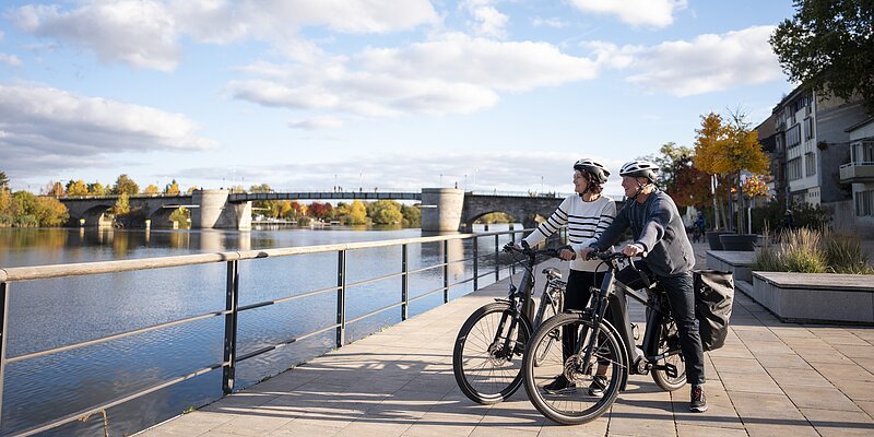 Ein älteres Pärchen steht mit ihren E-Bikes am Main in Kitzingen und genießt die Aussicht auf den Main und die Steinbrücke